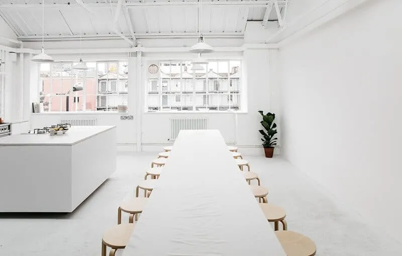 Salle blanche épurée avec long table et chaises en bois autour; espace lumineux prêt pour réunion, cuisine à gauche. 14 choses à jeter en un jour pour être mieux
